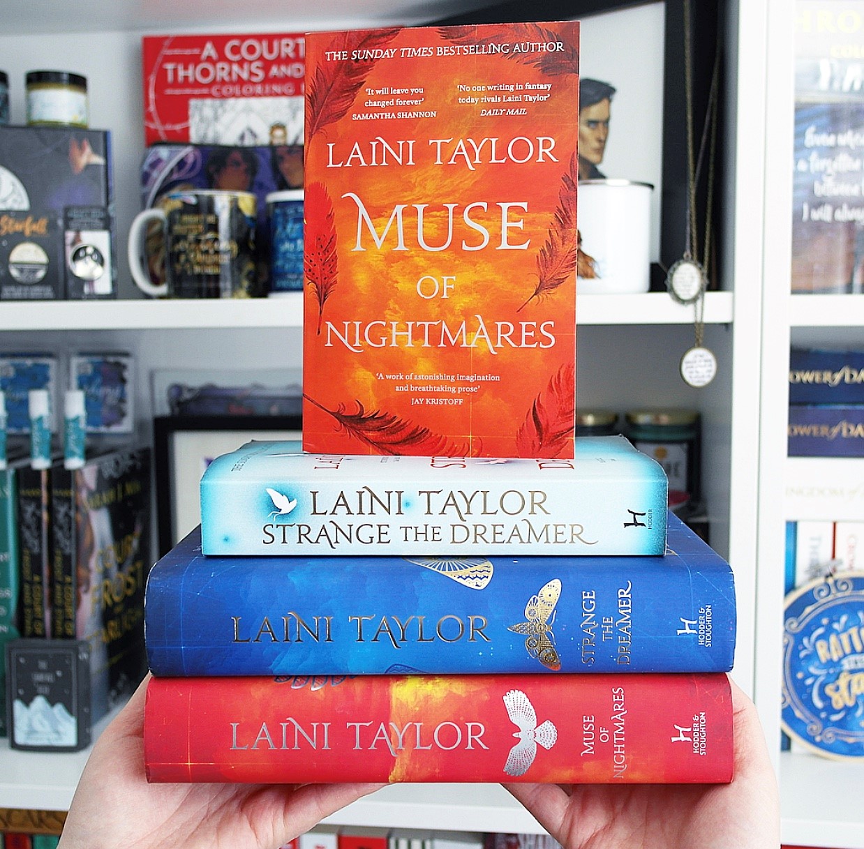 A stack of books by Laini Taylor. In order from top to bottom there is the red paperback edition of Muse of Nightmares stood on top of the blue paperback edition of Strange the Dreamer, the blue hardback edition of Strange the Dreamer and the red hardback edition of Muse of Nightmares. The last three books are stacked horizontally on top of one another.